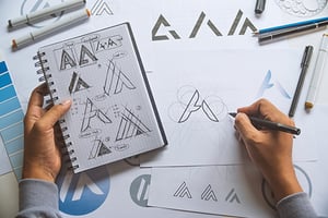 Researching your logo design will set you up for success. Hands sketch different versions of the letter "A" for a logo.