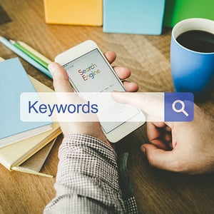 Keywords are vital in SEO strategy. “Keywords” in a translucent search bar; hands hold a phone at a desk with coffee cup.