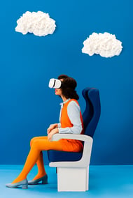 Virtual reality can greatly benefit travel industry websites. A women wears a VR headset, sits in an airplane chair with clouds in background. 
